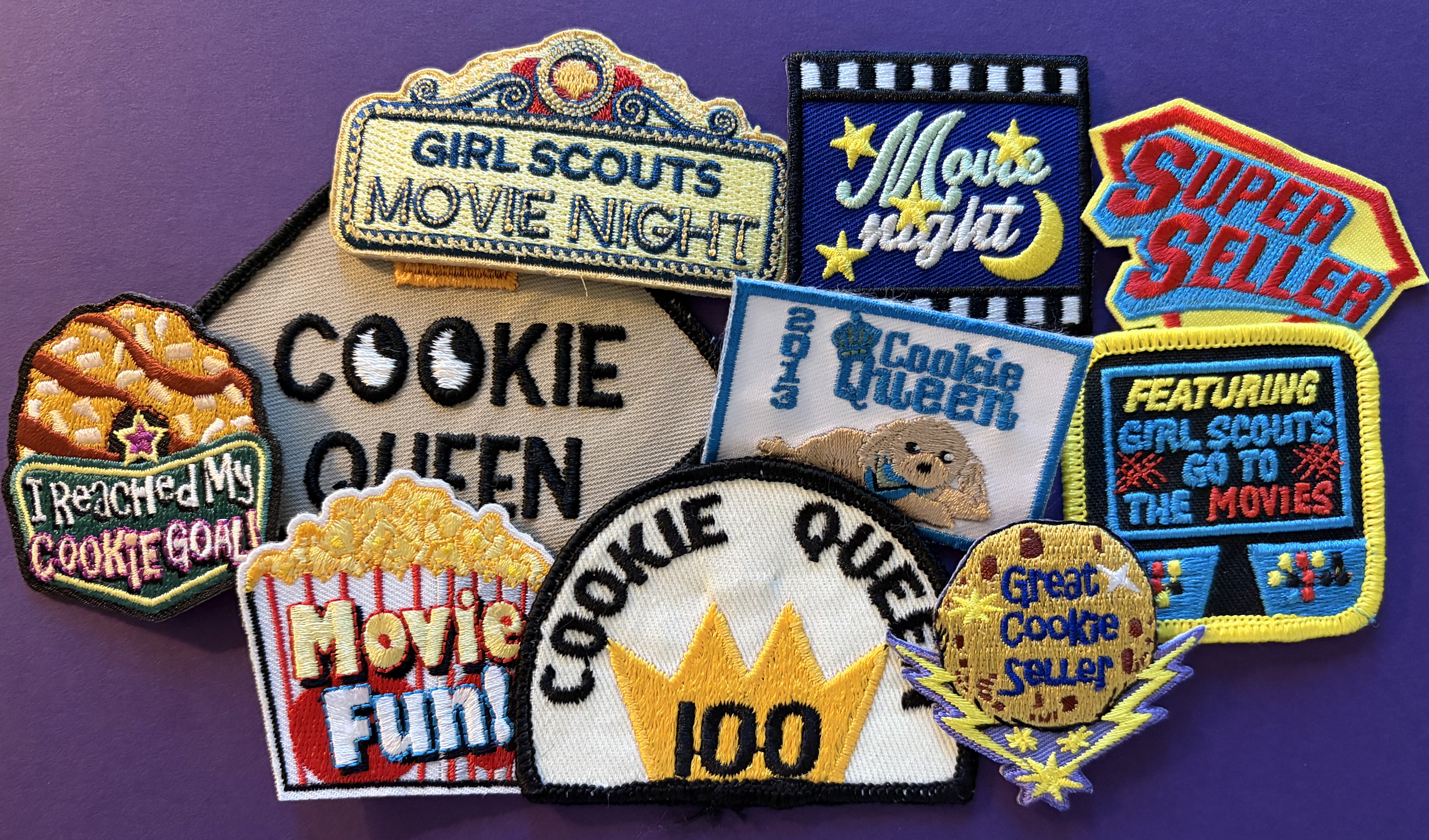 A collection of colorful Girl Scout patches on a purple background. Patches include phrases like Cookie Queen, Super Seller, Movie Night, and Great Cookie Seller, celebrating achievements in selling Girl Scout cookies, with popcorn and stars adorning some.